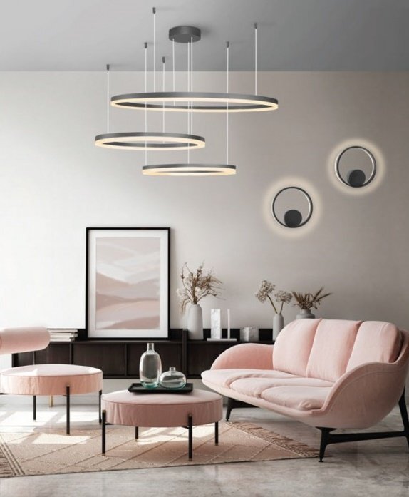 W nowoczesnym salonie znajduje się lampa wisząca z trzema okrągłymi pierścieniami. Wnętrze jest urządzone w jasnych kolorach z różową sofą i stolikiem kawowym.