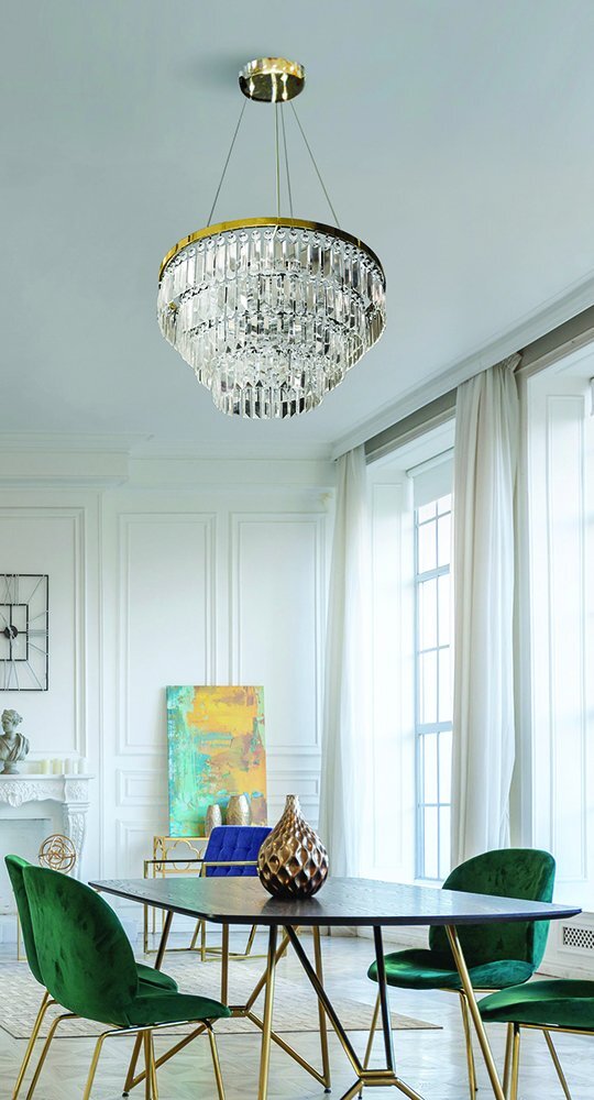 Elegancka lampa wisząca z kryształowymi elementami zawieszona w nowoczesnym wnętrzu z dużymi oknami i zielonymi krzesłami przy okrągłym stole. W tle widoczny jest kolorowy obraz oparty o ścianę.