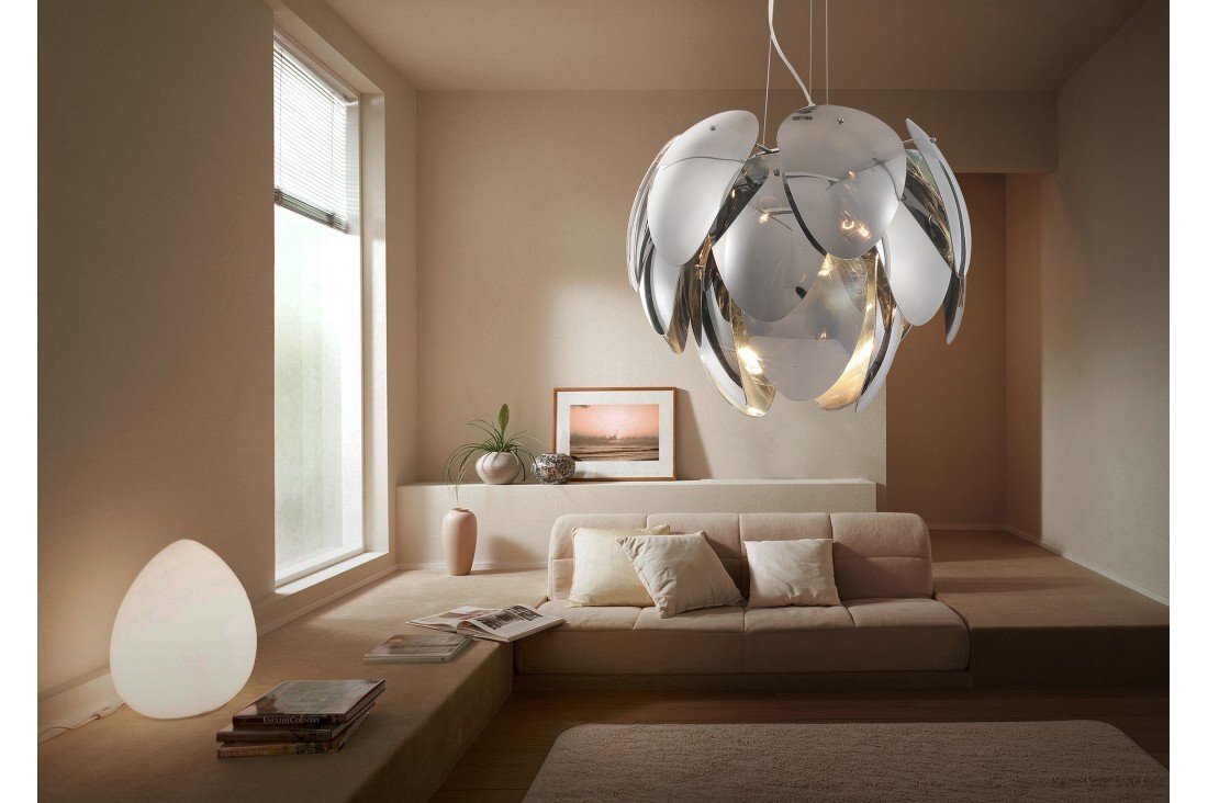 W nowoczesnym wnętrzu znajduje się lampa wisząca o futurystycznym designie, przypominająca kwiat z metalowymi płatkami. W pomieszczeniu jest także sofa z poduszkami, stolik z książkami i dekoracje w postaci roślin oraz obrazów.
