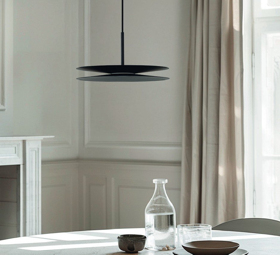 Elegancka, czarna lampa wisząca o nowoczesnym designie zawieszona nad stołem. Na stole znajdują się szklana butelka, filiżanka i miska.