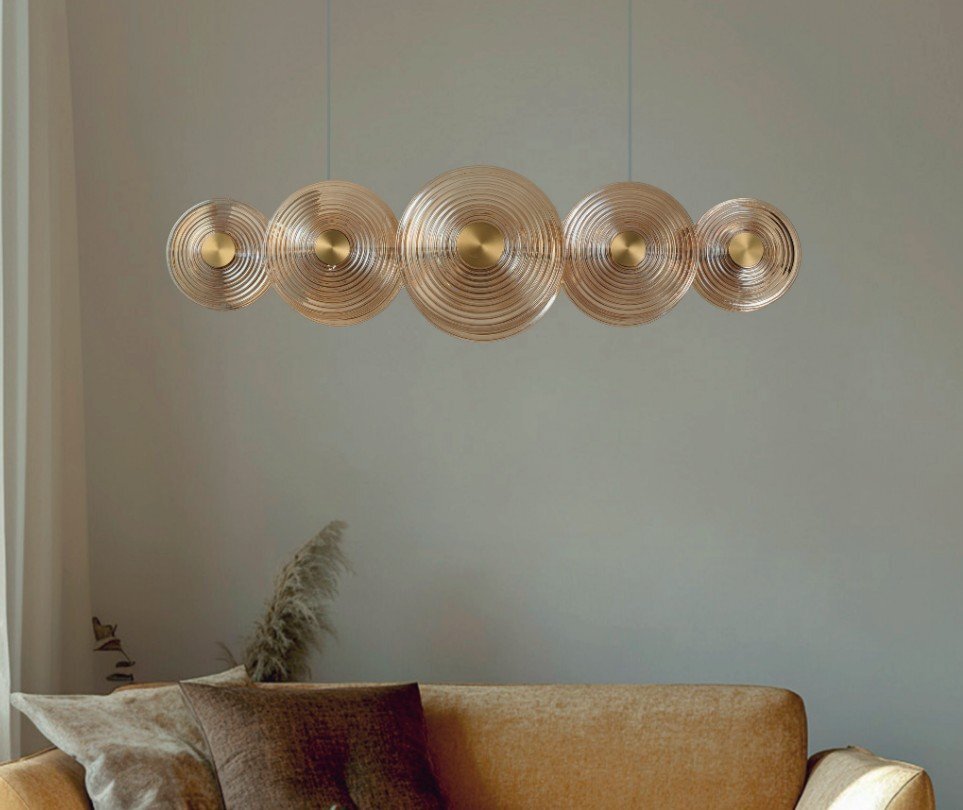 Na obrazie widoczna jest lampa wisząca z pięcioma okrągłymi kloszami o koncentrycznych wzorach, zawieszona nad sofą w salonie. W tle znajduje się jasna ściana i zasłona.