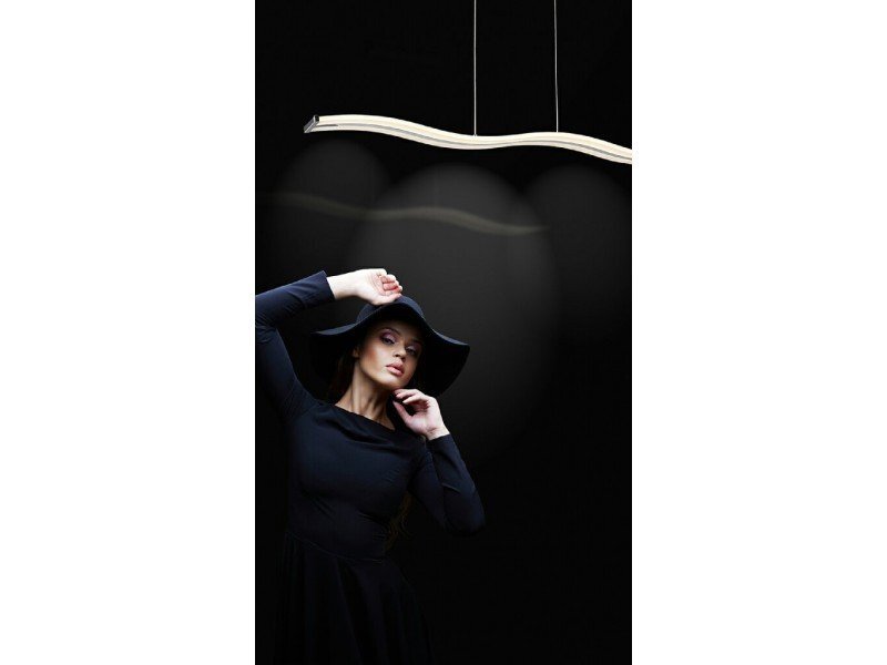 Na obrazie widoczna jest lampa wisząca o falistym kształcie, zawieszona na cienkich linkach. W tle znajduje się osoba ubrana na czarno, pozująca na ciemnym tle.