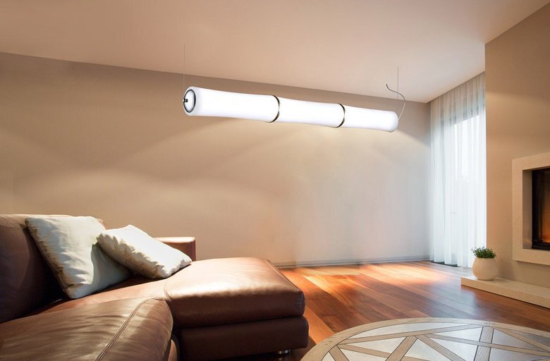 Wnętrze pokoju z nowoczesną lampą wiszącą o cylindrycznym kształcie, emitującą jasne światło. W tle znajduje się skórzana sofa z poduszkami oraz kominek, a na podłodze leży dywan na drewnianych panelach.