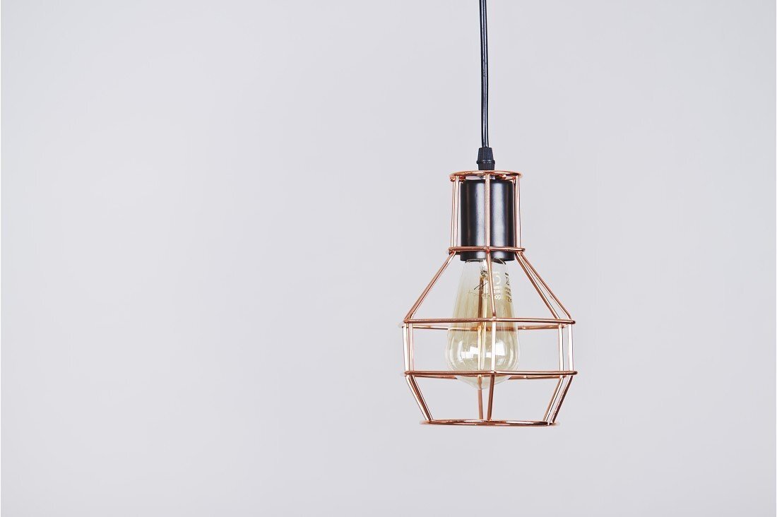 Lampa wisząca z metalowym kloszem w kształcie klatki, w kolorze miedzianym, z widoczną żarówką wewnątrz. Zawieszona na czarnym przewodzie na tle jasnej ściany.