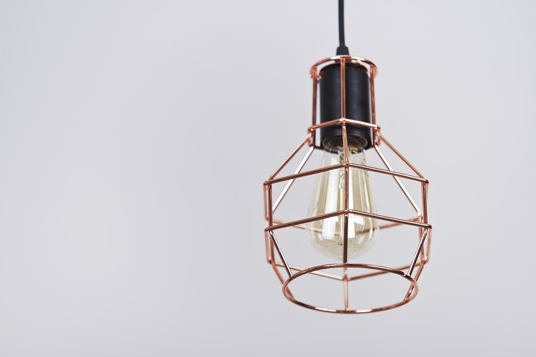 Lampa wisząca z metalowym kloszem w kształcie geometrycznej klatki, w której znajduje się żarówka. Klosz ma miedziany kolor, a przewód jest czarny.