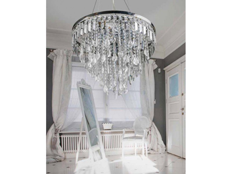 Na obrazie widoczna jest elegancka lampa wisząca z kryształowymi elementami, zawieszona w jasnym pomieszczeniu z dużym oknem i białymi zasłonami. W tle znajduje się lustro na stojaku oraz białe krzesło przy kaloryferze.