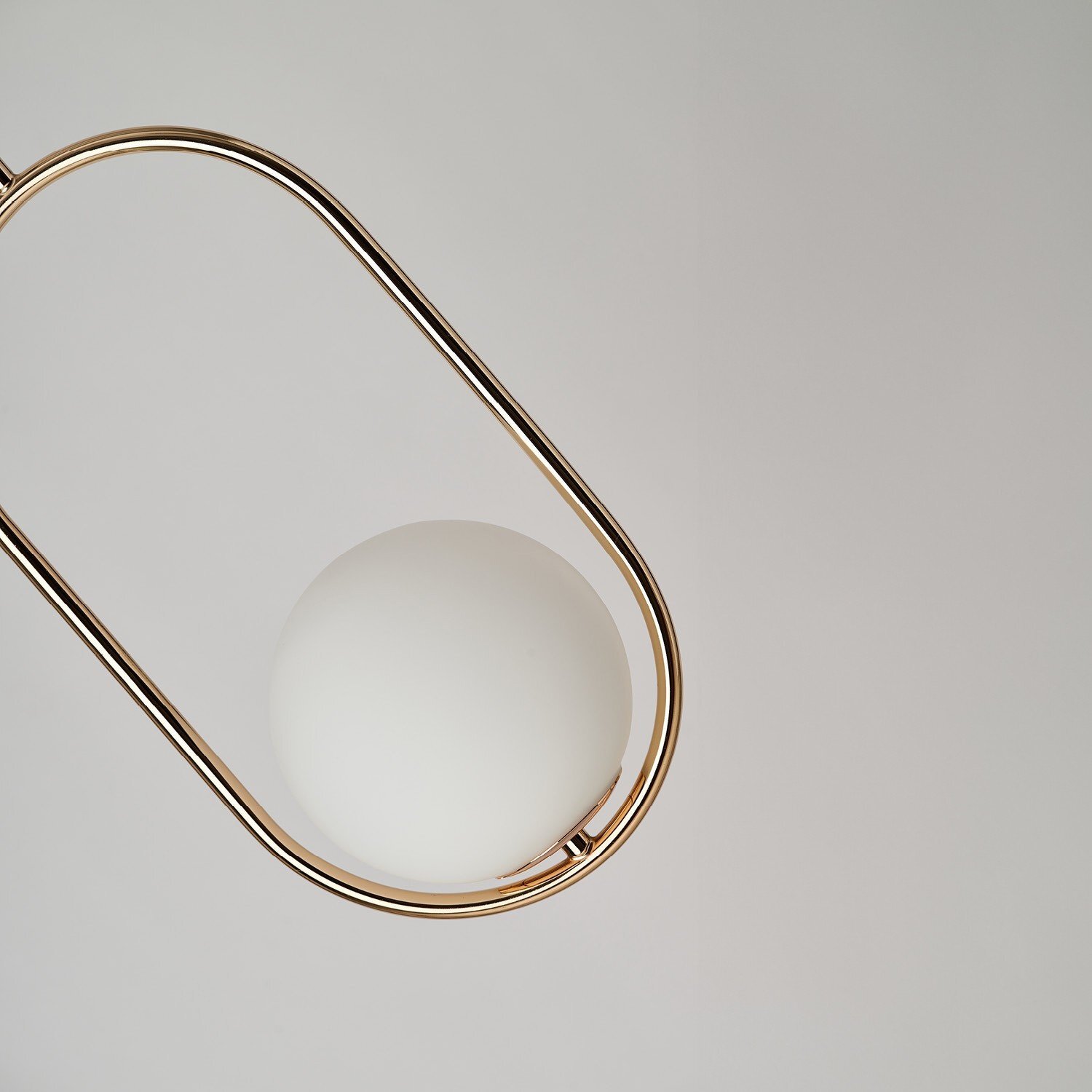 Na obrazie widoczna jest nowoczesna lampa wisząca z białym, kulistym kloszem otoczonym metalową, złotą ramą w kształcie elipsy. Tło jest jednolite i jasne.