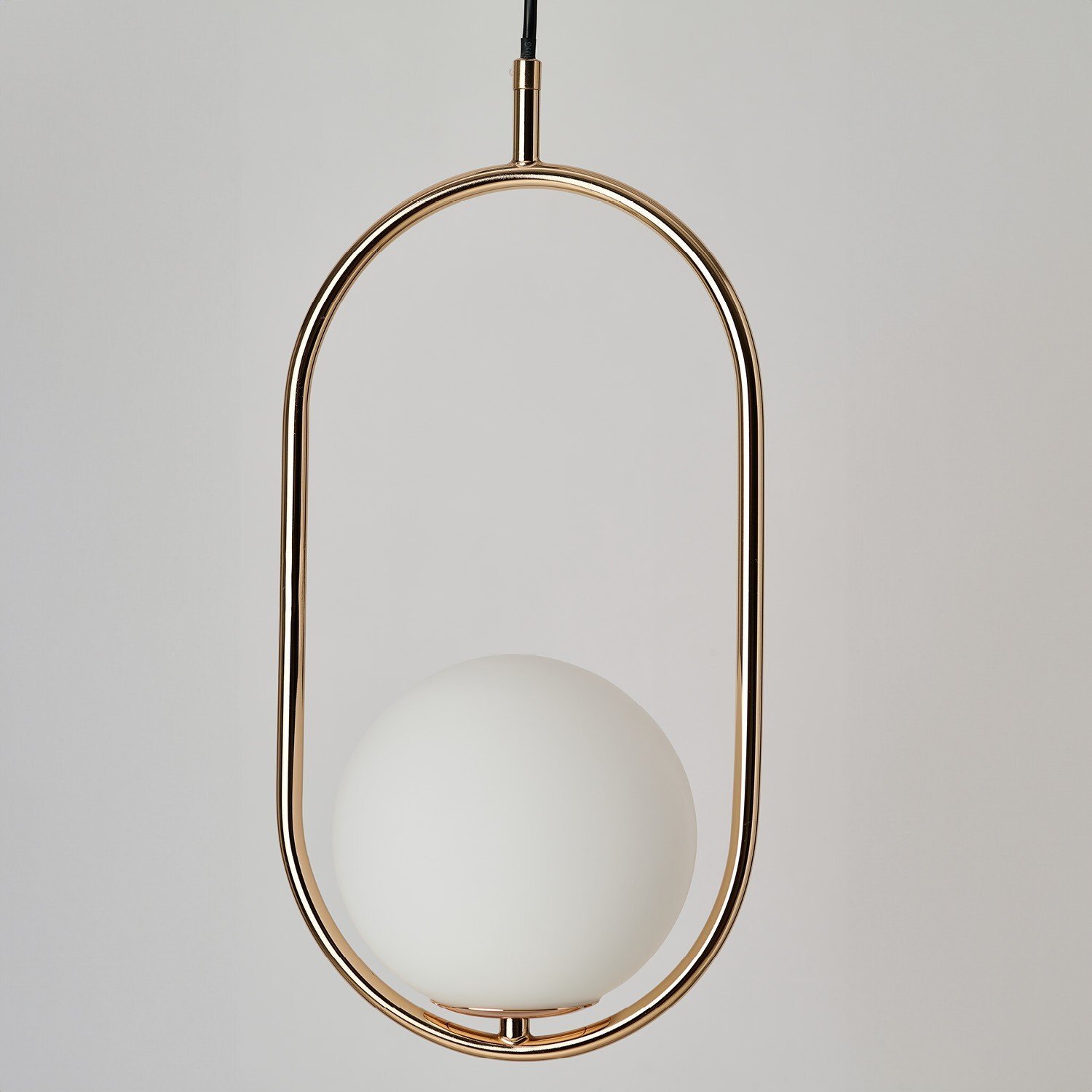 Na obrazie widoczna jest nowoczesna lampa wisząca z metalowym, złotym stelażem w kształcie owalu i białym, kulistym kloszem. Lampa zawieszona jest na czarnym przewodzie.