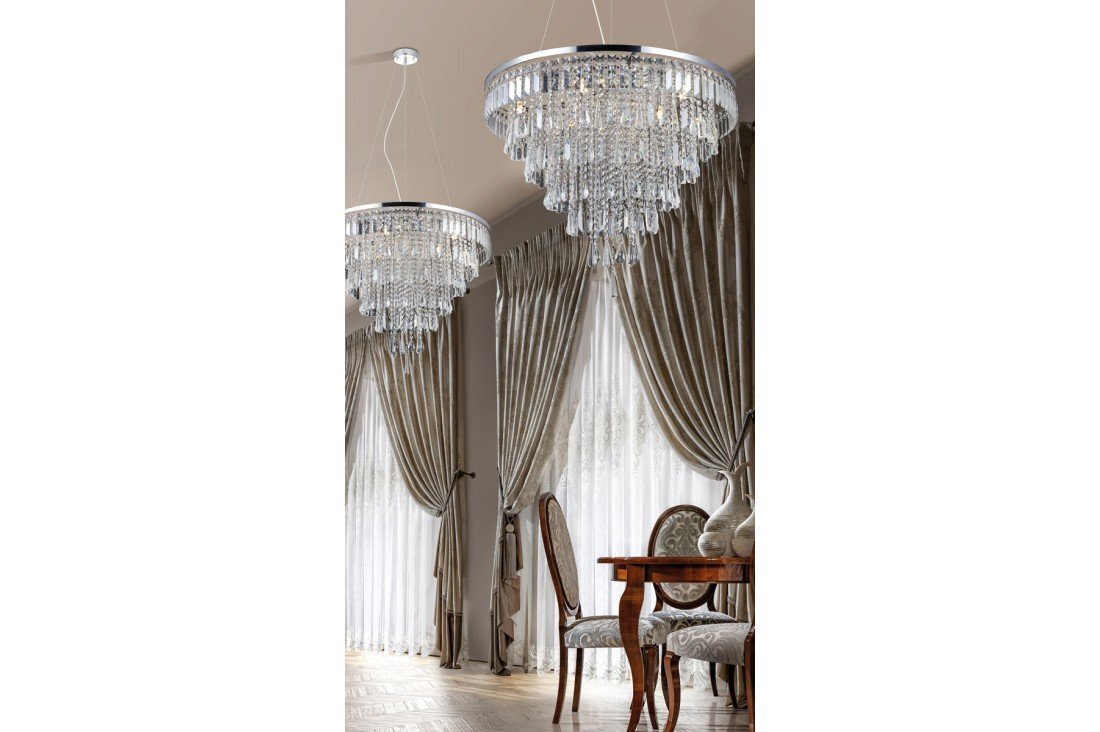 Na obrazie widoczne są dwie eleganckie, kryształowe lampy wiszące w pomieszczeniu z zasłonami i drewnianym stołem z krzesłami. W tle znajdują się duże okna z zasłonami w kolorze beżowym.