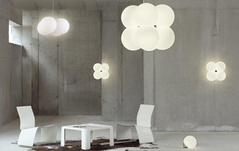 W nowoczesnym wnętrzu z betonowymi ścianami znajdują się białe, minimalistyczne meble oraz kilka lamp o kształcie przypominającym kwiaty. Lampy emitują miękkie, rozproszone światło, tworząc przytulną atmosferę.
