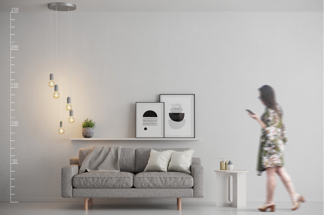 Wnętrze pokoju z szarą sofą, nad którą wisi lampa z pięcioma żarówkami. Obok sofy znajduje się półka z rośliną i dwoma obrazami, a po prawej stronie stoi stolik z filiżanką.