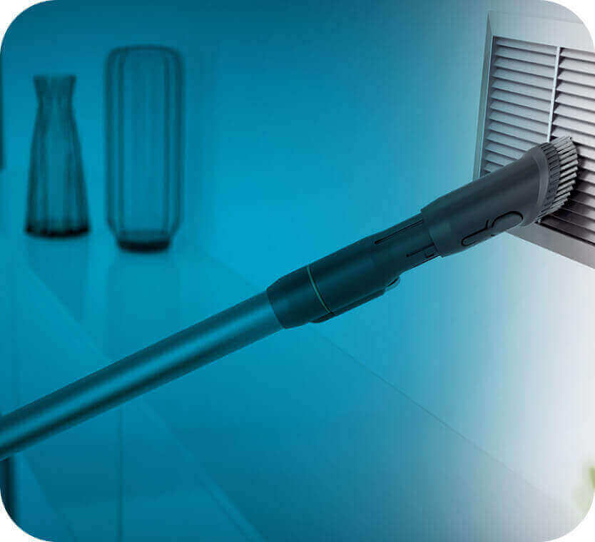 Na obrazie widoczny jest odkurzacz z długą rurą, który czyści kratkę wentylacyjną. W tle znajdują się dwa wazony na półce.