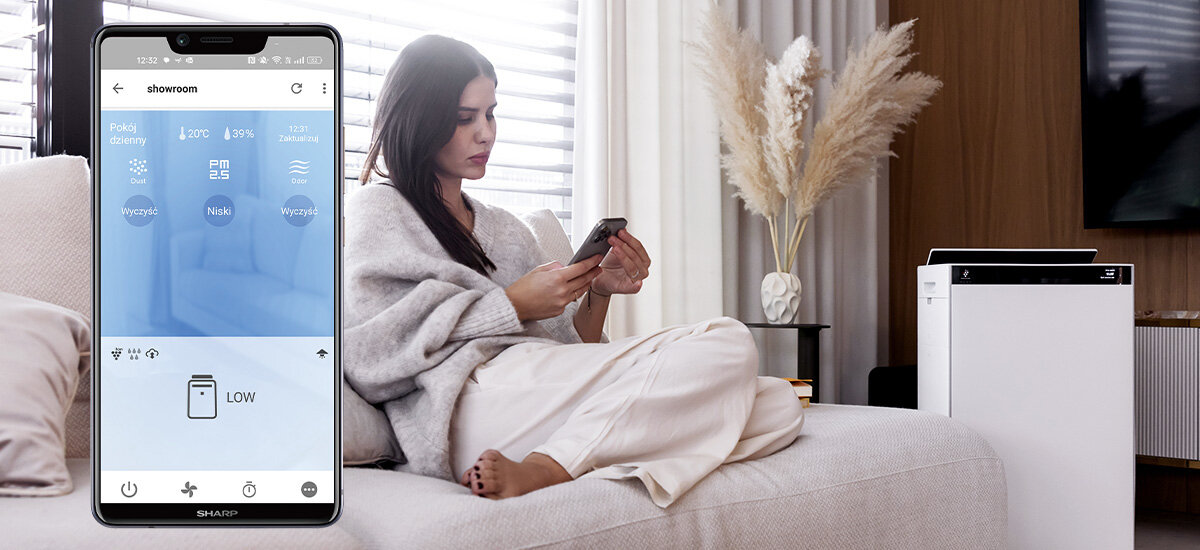 Kobieta siedzi na kanapie z telefonem w ręku, obok niej stoi oczyszczacz powietrza SHARP. Na ekranie telefonu widoczna jest aplikacja z napisem \'showroom\' i ikonami dotyczącymi sterowania urządzeniem.