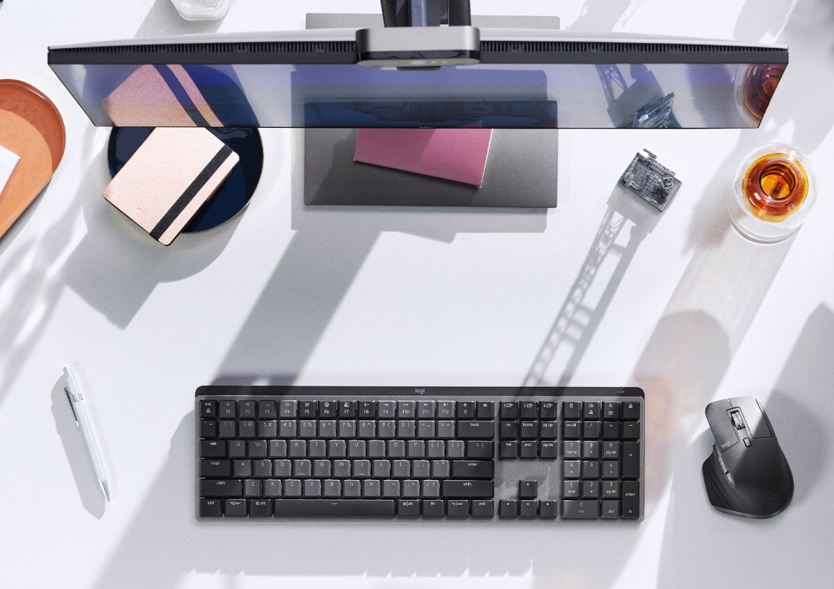 Na biurku znajduje się klawiatura mechaniczna z ciemnymi klawiszami, obok której leży mysz komputerowa. W tle widoczny jest monitor, notatnik, długopis oraz szklanka z napojem.