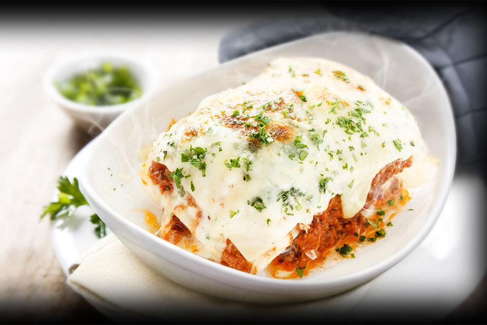 Na obrazie widoczna jest porcja lasagne w białym naczyniu, pokryta roztopionym serem i posypana świeżymi ziołami. W tle znajduje się niewielka miseczka z zielonymi ziołami.