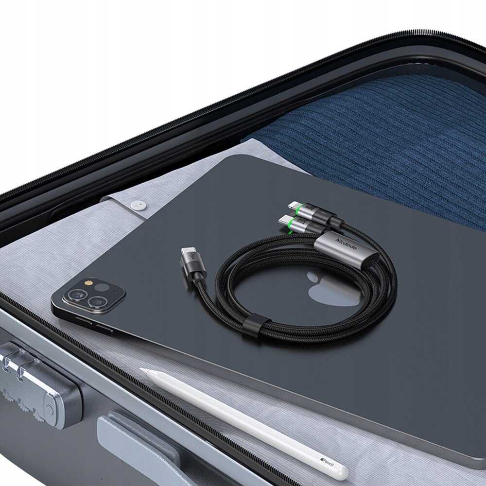 Na obrazie widoczny jest czarny kabel USB-C z dwoma końcówkami, leżący na ciemnym laptopie z logo w kształcie jabłka. W tle znajduje się walizka z ubraniami i białym rysikiem.