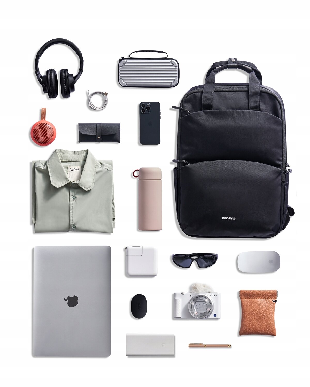 Na obrazie znajduje się czarny plecak z logo \'innostyle\', otoczony różnymi przedmiotami, takimi jak laptop, słuchawki, telefon, aparat, okulary przeciwsłoneczne, koszula, termos i inne akcesoria. Wszystkie przedmioty są starannie ułożone na białym tle.