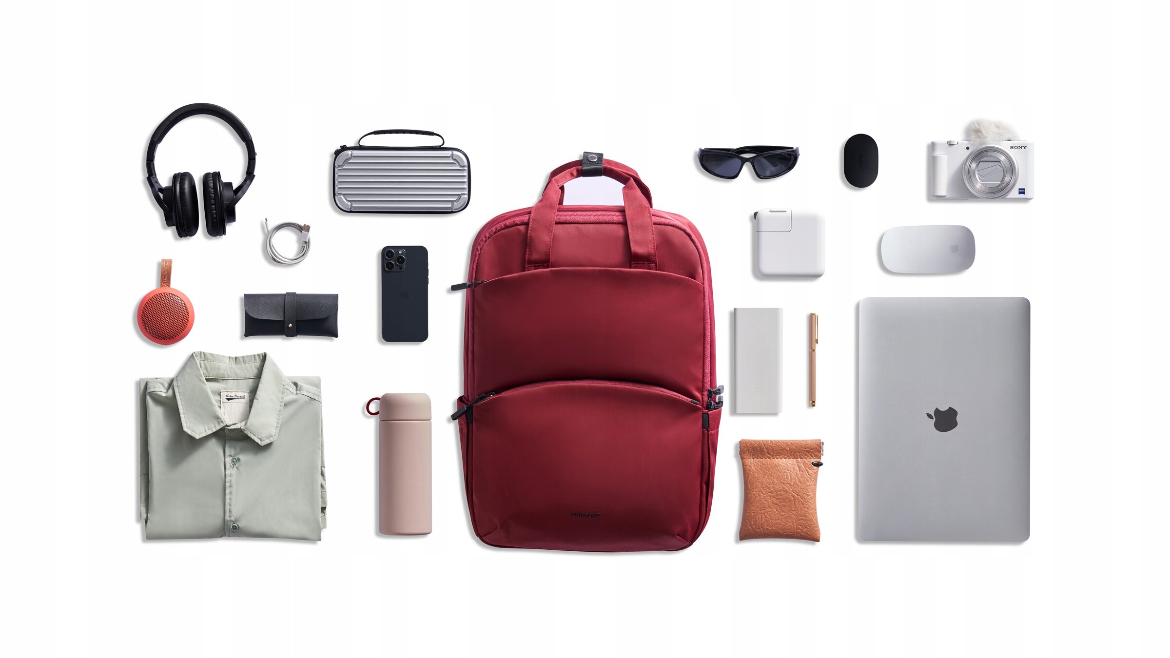 Na obrazie widoczny jest czerwony plecak otoczony różnymi przedmiotami, takimi jak słuchawki, laptop, aparat fotograficzny, okulary przeciwsłoneczne i inne akcesoria. Plecak ma prosty design z dwoma uchwytami na górze.