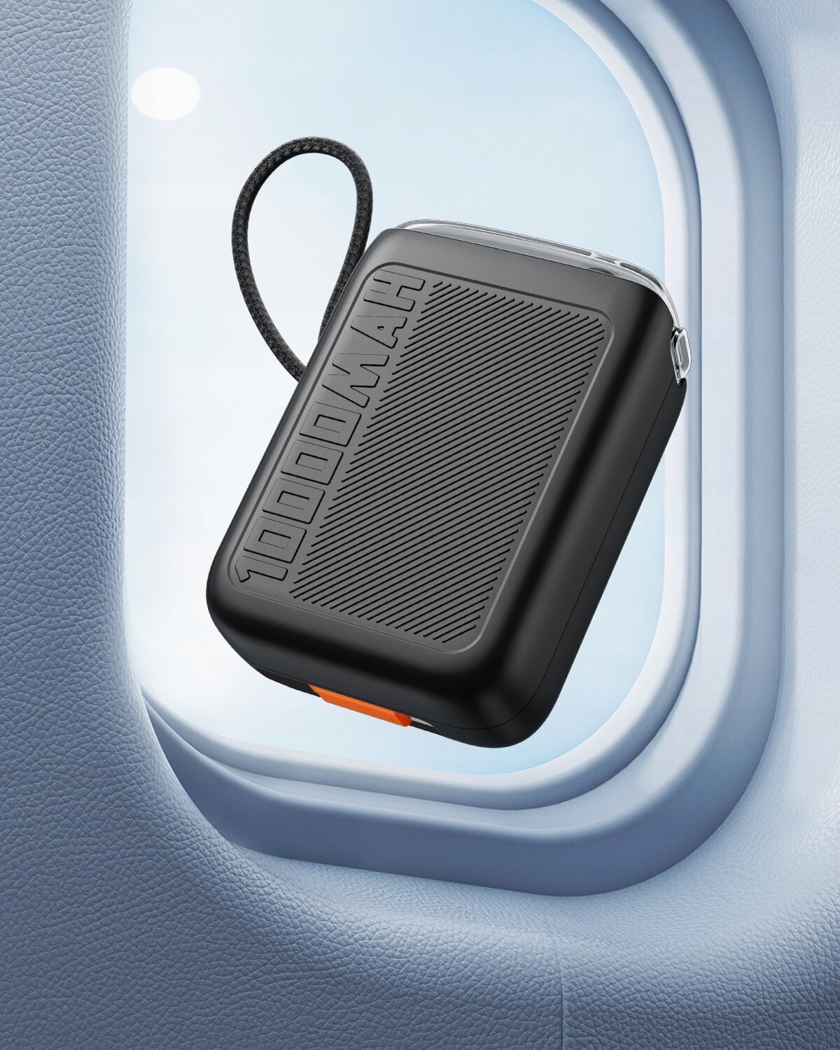 Czarny powerbank z napisem \'10000MAH\' widoczny jest na tle okna samolotu. Posiada teksturowaną powierzchnię i krótki uchwyt z plecionego materiału.