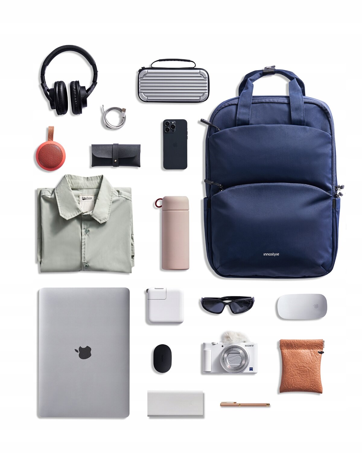 Na obrazie widoczny jest niebieski plecak z logo \'innostyle\', otoczony różnymi przedmiotami, takimi jak laptop, słuchawki, aparat fotograficzny, okulary przeciwsłoneczne, koszula, termos i inne akcesoria elektroniczne.