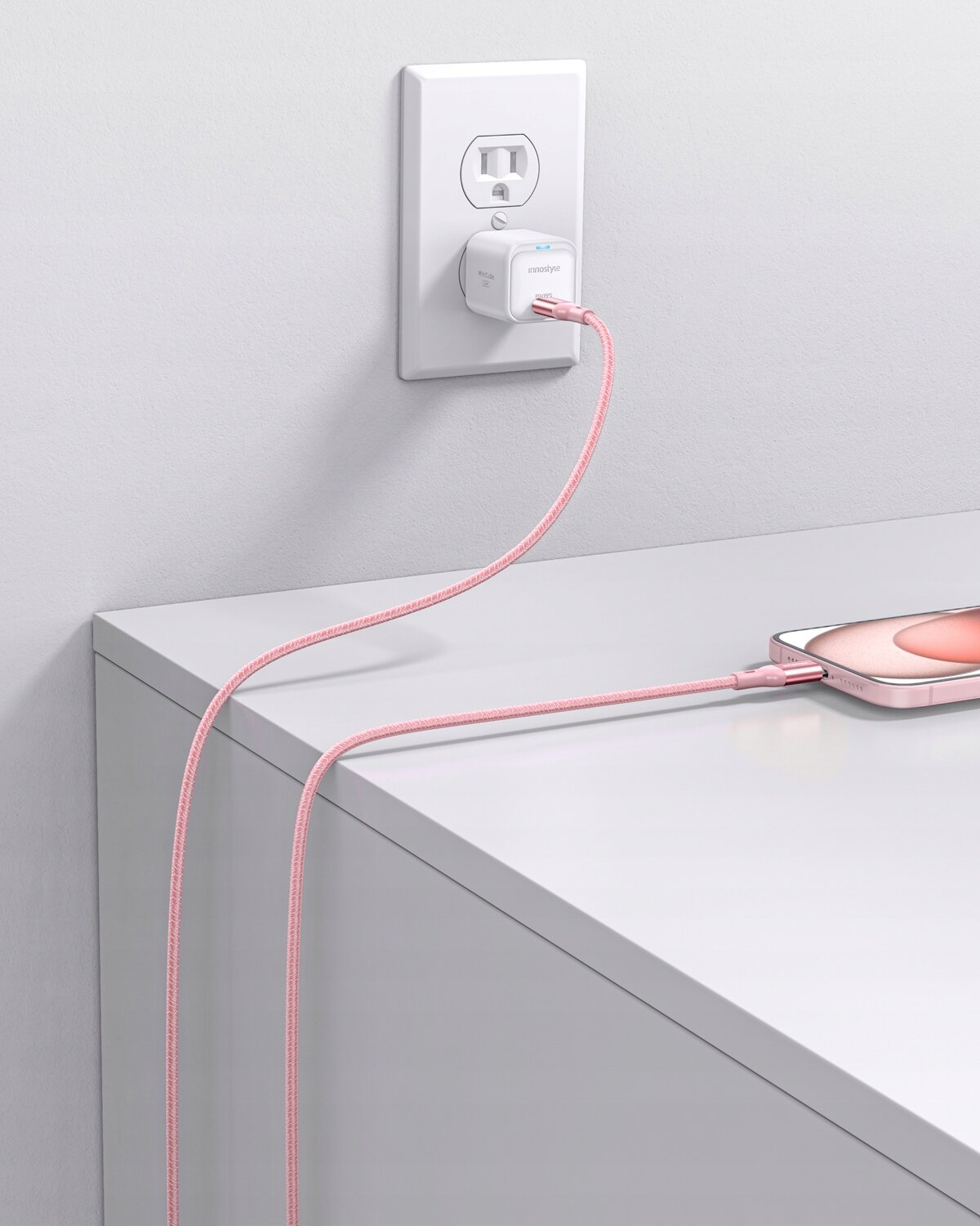 Różowy kabel USB-C podłączony do białej ładowarki w gniazdku ściennym, z drugiej strony podłączony do smartfona leżącego na blacie. Na ładowarce widoczne jest logo \'Innostyle\'.