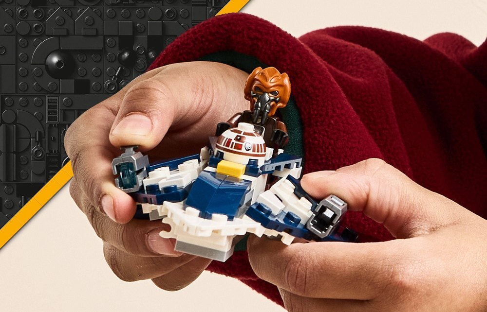 Osoba trzyma w dłoniach mały model statku kosmicznego z klocków LEGO, z figurką postaci w hełmie. Tło jest ozdobione wzorem z klocków LEGO w kolorze czarnym z żółtym paskiem.