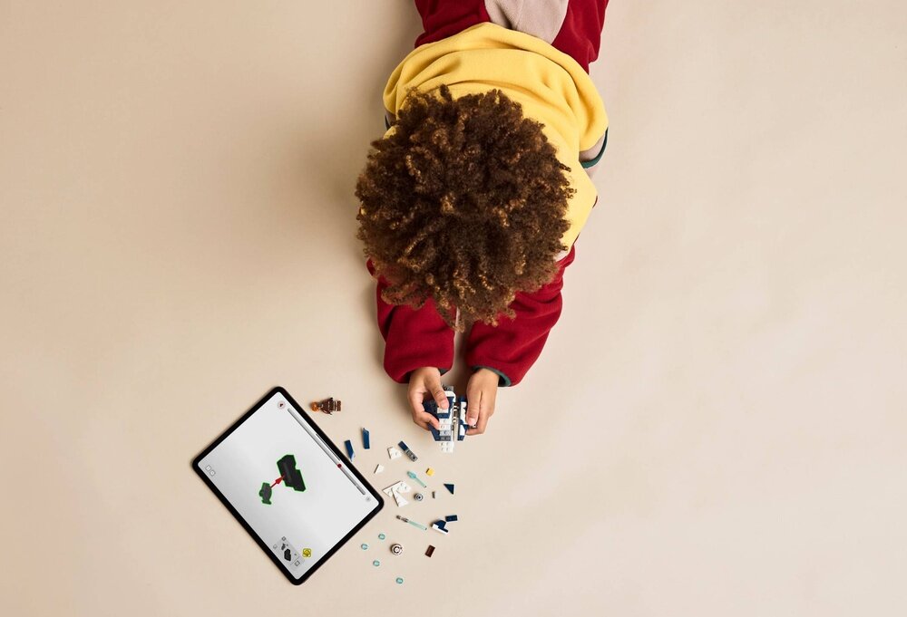Dziecko w żółtej bluzie bawi się klockami LEGO, obok leży tablet z instrukcją budowy. Na podłodze rozłożone są różnokolorowe elementy LEGO.