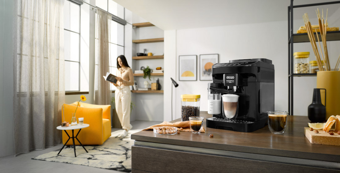 Na blacie kuchennym stoi czarny ekspres do kawy z logo \'DeLonghi Magnifica Evo\', obok filiżanka z kawą i szklanka z mlekiem. W tle widoczna jest osoba czytająca książkę w jasnym, nowoczesnym wnętrzu z żółtymi akcentami.