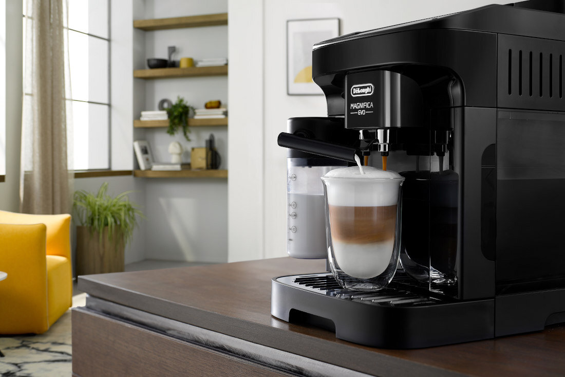Czarny ekspres do kawy z logo \'DELONGHI Magnifica Evo\' przygotowuje kawę z mleczną pianką w szklance. W tle widoczne są półki z dekoracjami i żółty fotel.