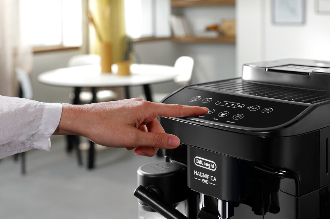 Osoba dotyka panelu sterowania czarnego ekspresu do kawy z widocznym logo \'DeLonghi Magnifica Evo\'. W tle znajduje się rozmyty widok wnętrza z okrągłym stołem i krzesłami.