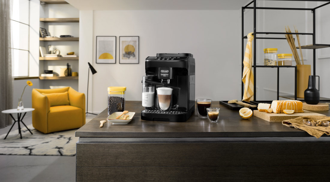 Na blacie kuchennym stoi czarny ekspres do kawy z logo DELONGHI, obok filiżanki z kawą i talerza z pieczywem. W tle widoczny jest żółty fotel oraz półki z dekoracjami.