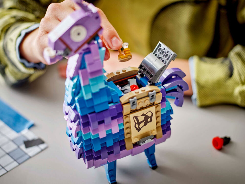 Na obrazie widoczny jest model LEGO przedstawiający kolorową lamę z otwartym schowkiem, z którego wyciągana jest mała figurka. W tle widać rękę osoby manipulującej elementami zestawu.