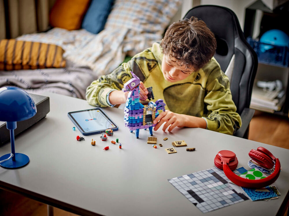 Na biurku znajduje się zestaw LEGO w kształcie kolorowej lamy, obok której leży tablet i kilka małych elementów LEGO. W tle widać łóżko z poduszkami oraz lampkę biurkową.