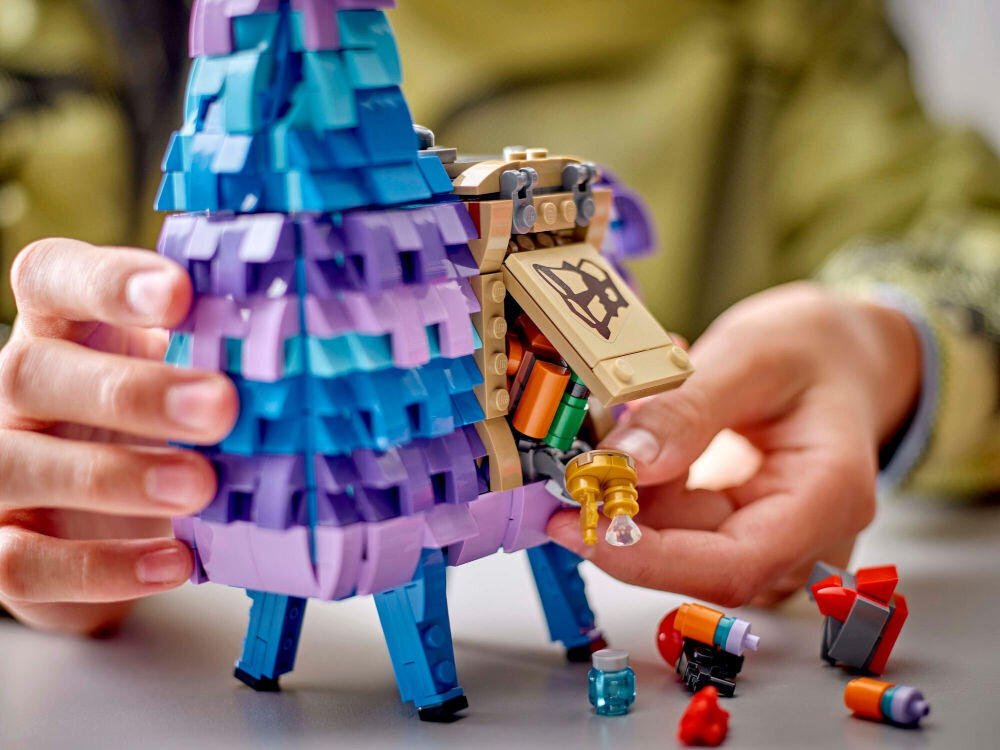 Osoba trzyma model LEGO przedstawiający kolorową lamę z otwartym schowkiem, z którego wystają różne elementy. Na stole obok znajdują się małe, kolorowe klocki LEGO.