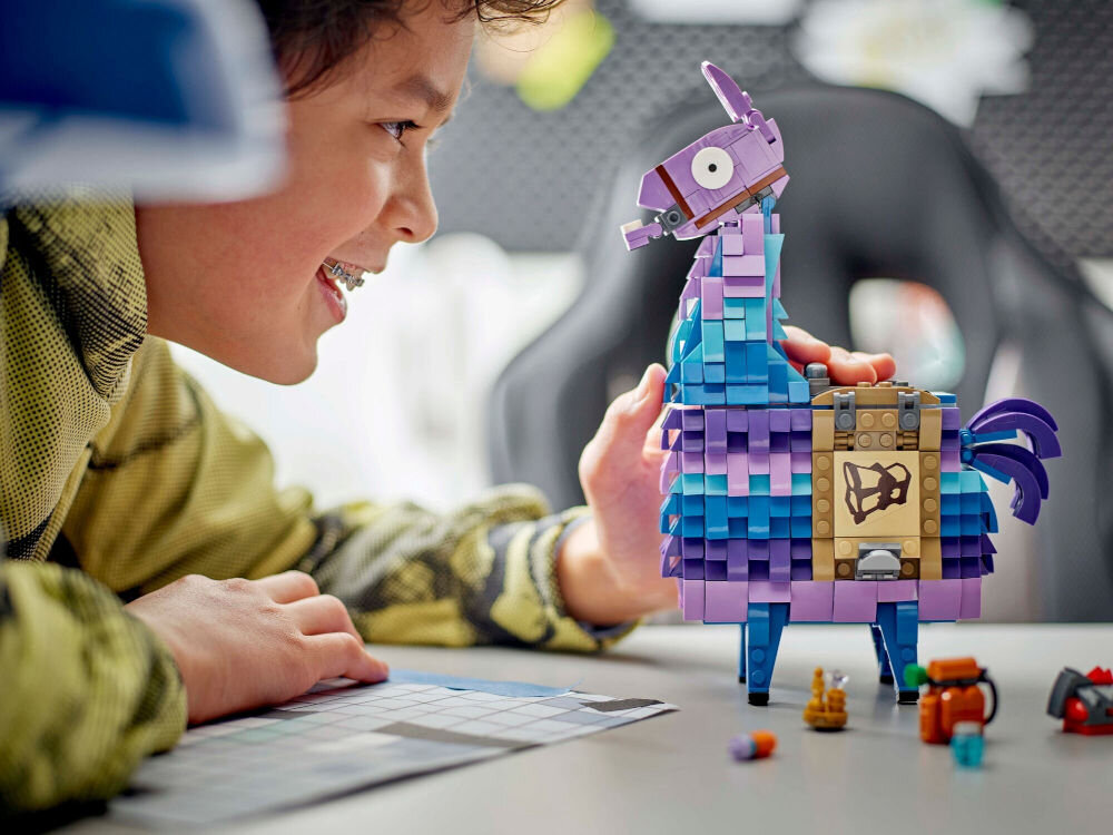 Na stole znajduje się kolorowy model lamy z klocków LEGO, obok którego leży kilka małych elementów. W tle widać osobę uśmiechającą się do modelu.