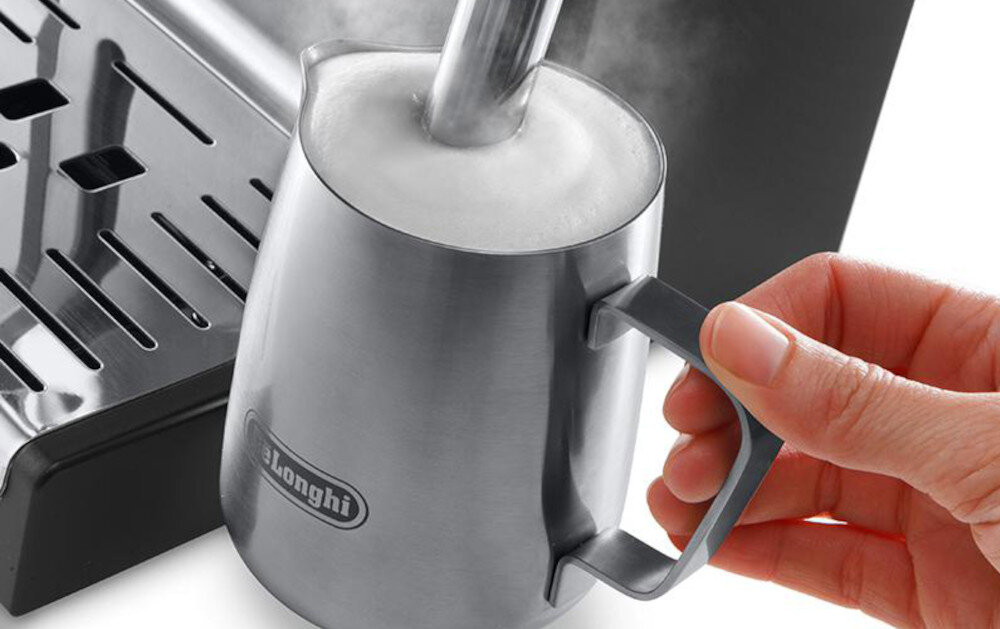 Osoba trzyma metalowy dzbanek z logo \'DeLonghi\', w którym spieniane jest mleko za pomocą dyszy parowej. Obok widoczna jest część ekspresu do kawy z metalową tacką ociekową.