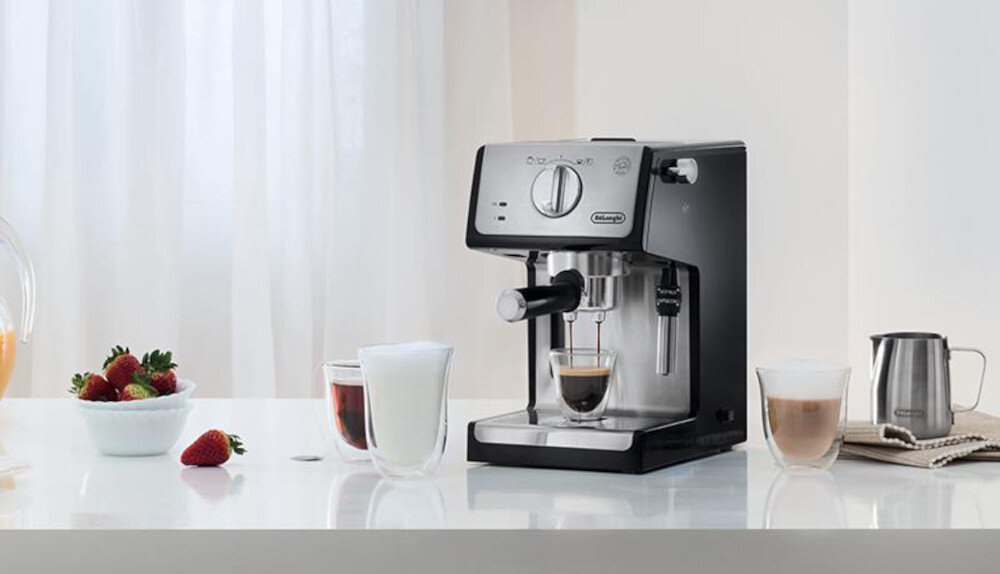 Na blacie stoi ekspres do kawy z logo \'DeLonghi\', obok którego znajdują się szklanki z różnymi napojami oraz miska z truskawkami.