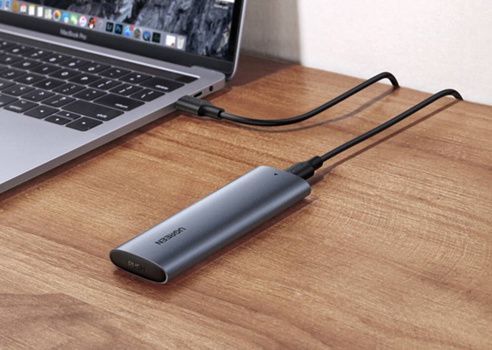 Na drewnianym blacie znajduje się laptop z podłączoną obudową dysku UGREEN CM400. Obudowa jest podłączona do laptopa za pomocą czarnego kabla USB-C.