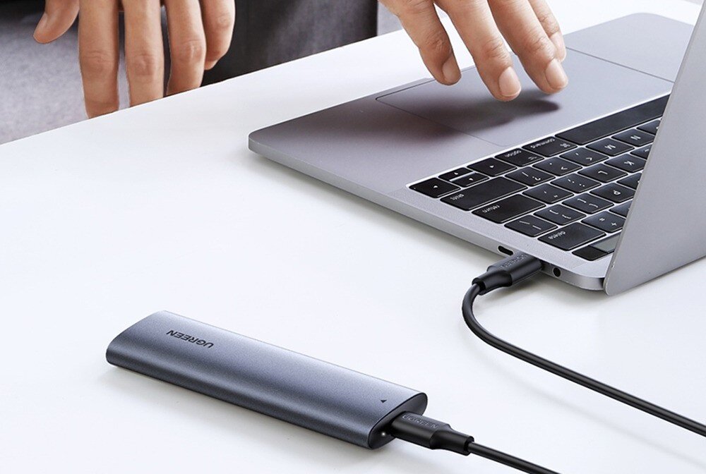 Na białym stole znajduje się laptop z otwartą klawiaturą, do którego podłączony jest szary, podłużny dysk zewnętrzny z napisem \'UGREEN\'. Obok laptopa widoczne są dłonie osoby, która go obsługuje.