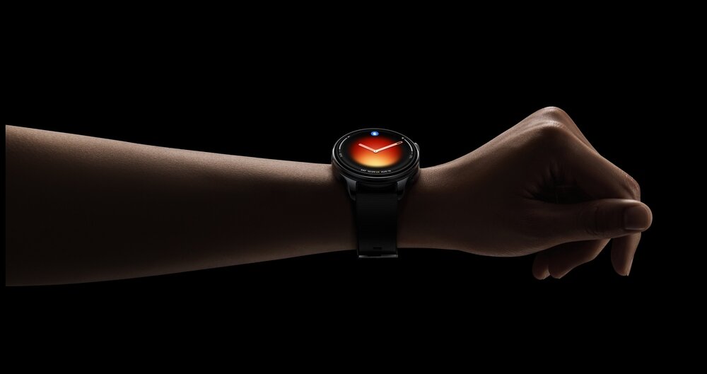 Na zdjęciu widoczny jest czarny smartwatch na nadgarstku, z okrągłym wyświetlaczem pokazującym tarczę zegara z czerwonym gradientem. Tło jest ciemne, co podkreśla kontrast urządzenia.