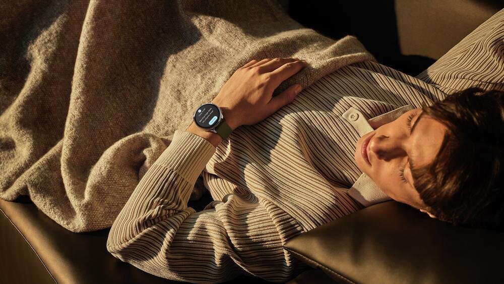 Osoba leży na kanapie, mając na nadgarstku czarny smartwatch z okrągłym wyświetlaczem. Smartwatch pokazuje ekran z tekstem, ale nie jest on czytelny.
