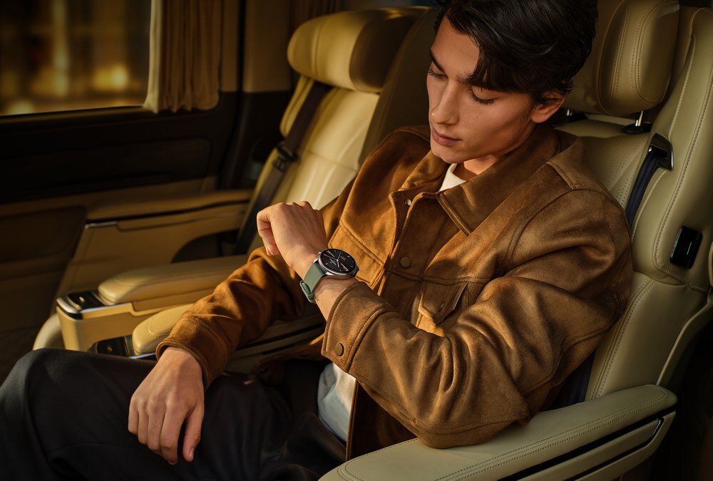 Osoba siedzi w samochodzie, patrząc na czarny smartwatch na nadgarstku. Wnętrze pojazdu jest eleganckie, z jasnymi skórzanymi siedzeniami.