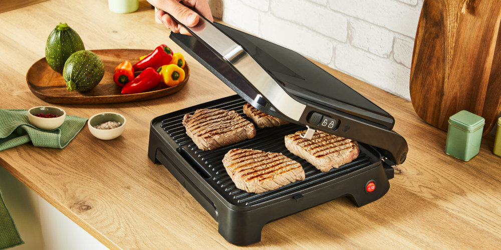 Na drewnianym blacie kuchennym znajduje się otwarty grill elektryczny z czterema kawałkami mięsa. Obok leżą warzywa, przyprawy i akcesoria kuchenne.