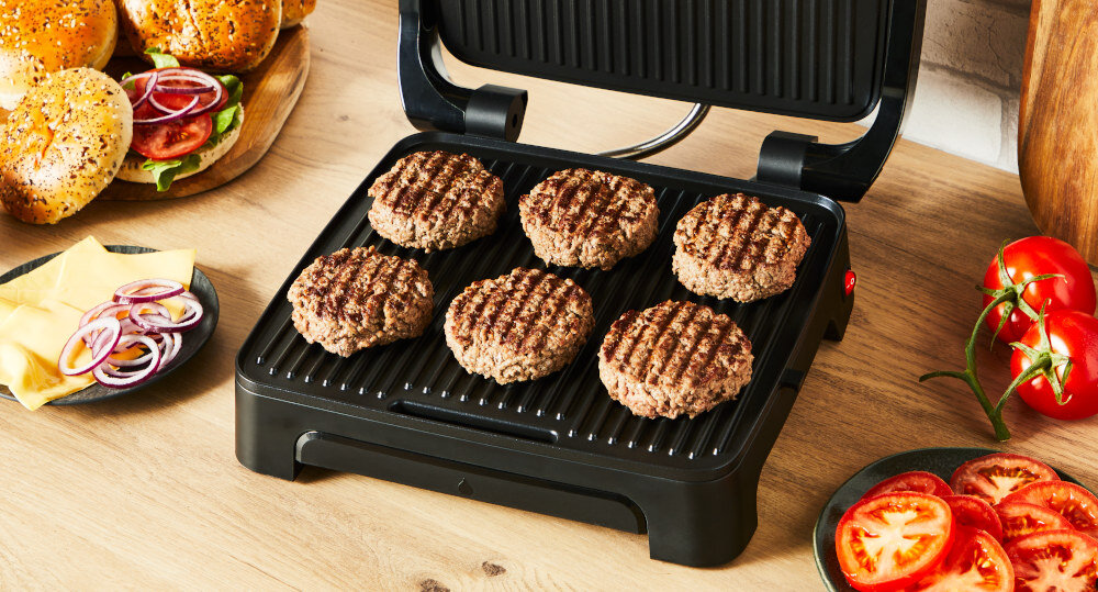 Na obrazie widoczny jest grill elektryczny z pięcioma grillowanymi burgerami na ruszcie. Obok znajdują się składniki do burgerów, takie jak bułki, ser, cebula i pomidory.