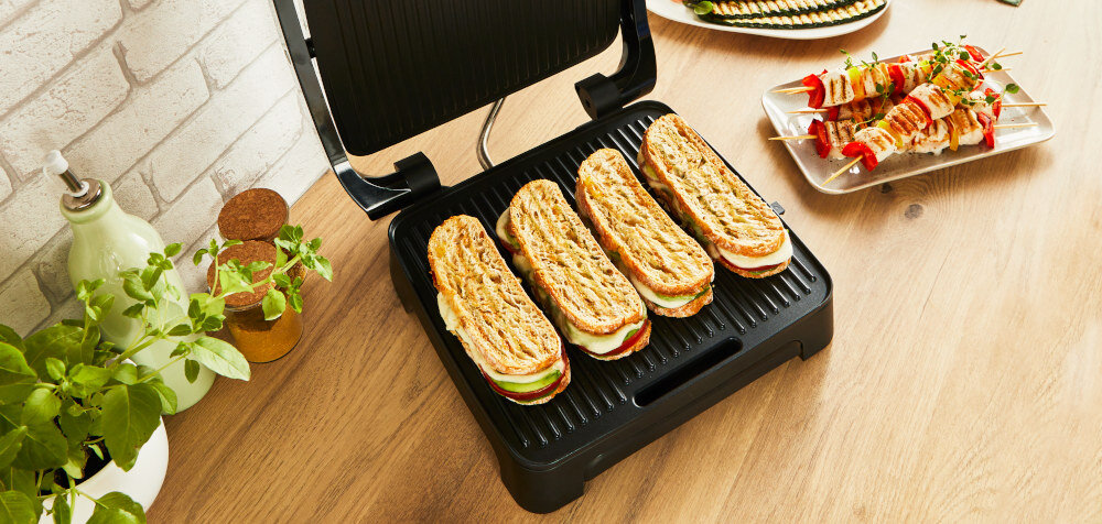 Na drewnianym blacie stoi otwarty grill elektryczny z czterema kanapkami w środku. Obok znajdują się zioła w doniczce i talerz z grillowanymi szaszłykami.