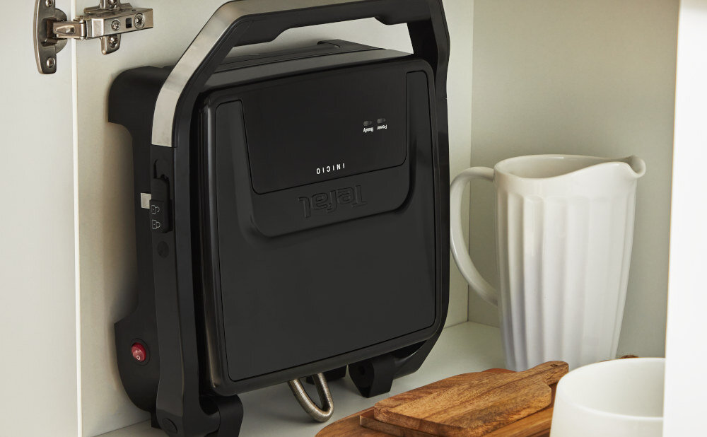 Czarny grill elektryczny z logo \'tefal\' stoi pionowo w szafce obok białego dzbanka i drewnianej deski. Na urządzeniu widoczny jest napis \'INICIO\'.