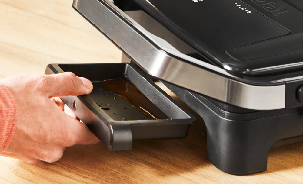 Osoba wyciąga tackę ociekową z elektrycznego grilla marki Tefal. Na grillu widoczne jest logo \'Tefal\' oraz napis \'INICIO\'.
