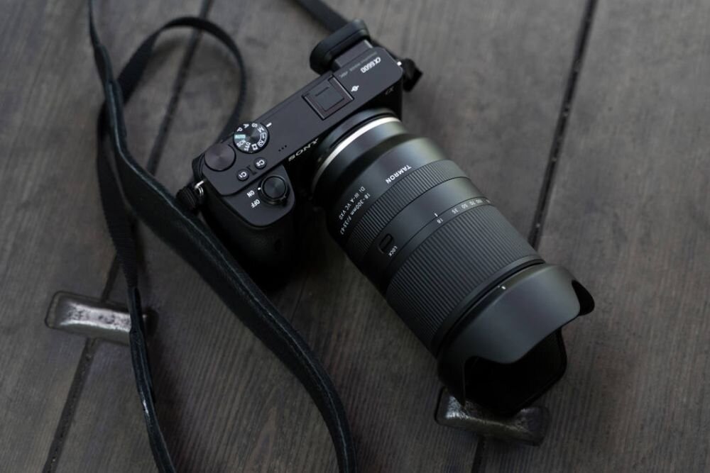 Na drewnianej powierzchni leży aparat fotograficzny z zamontowanym obiektywem TAMRON. Obiektyw ma widoczne oznaczenia i pierścienie do regulacji ostrości i zoomu.