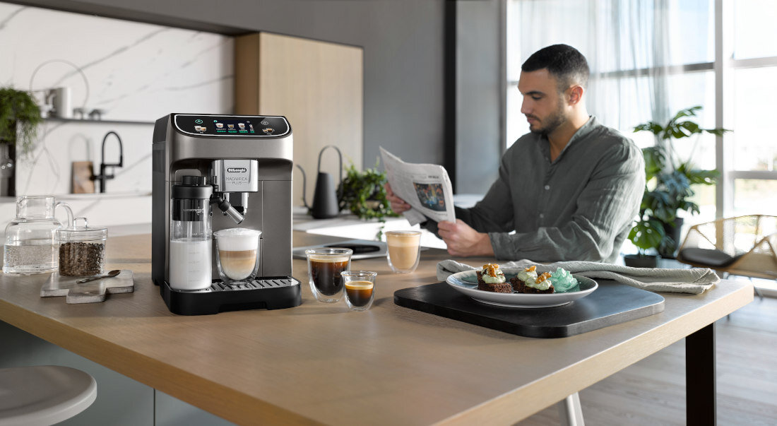 Na blacie kuchennym stoi ekspres do kawy z logo Delonghi, obok znajdują się filiżanki z kawą. W tle mężczyzna czyta gazetę przy stole z talerzem ciast.
