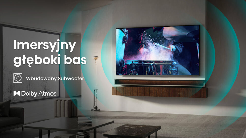 W salonie na ścianie wisi telewizor, na którym wyświetlany jest obraz perkusisty. Obok widoczny jest tekst: \'Imersyjny głęboki bas\', \'Wbudowany Subwoofer\', \'Dolby Atmos\'.